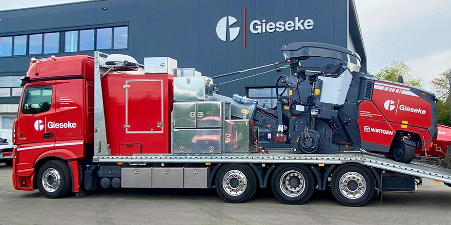 Eine Asphaltfräse, die transportiert wird vor einem grauen Gebäude mit der Aufschrift Gieseke.